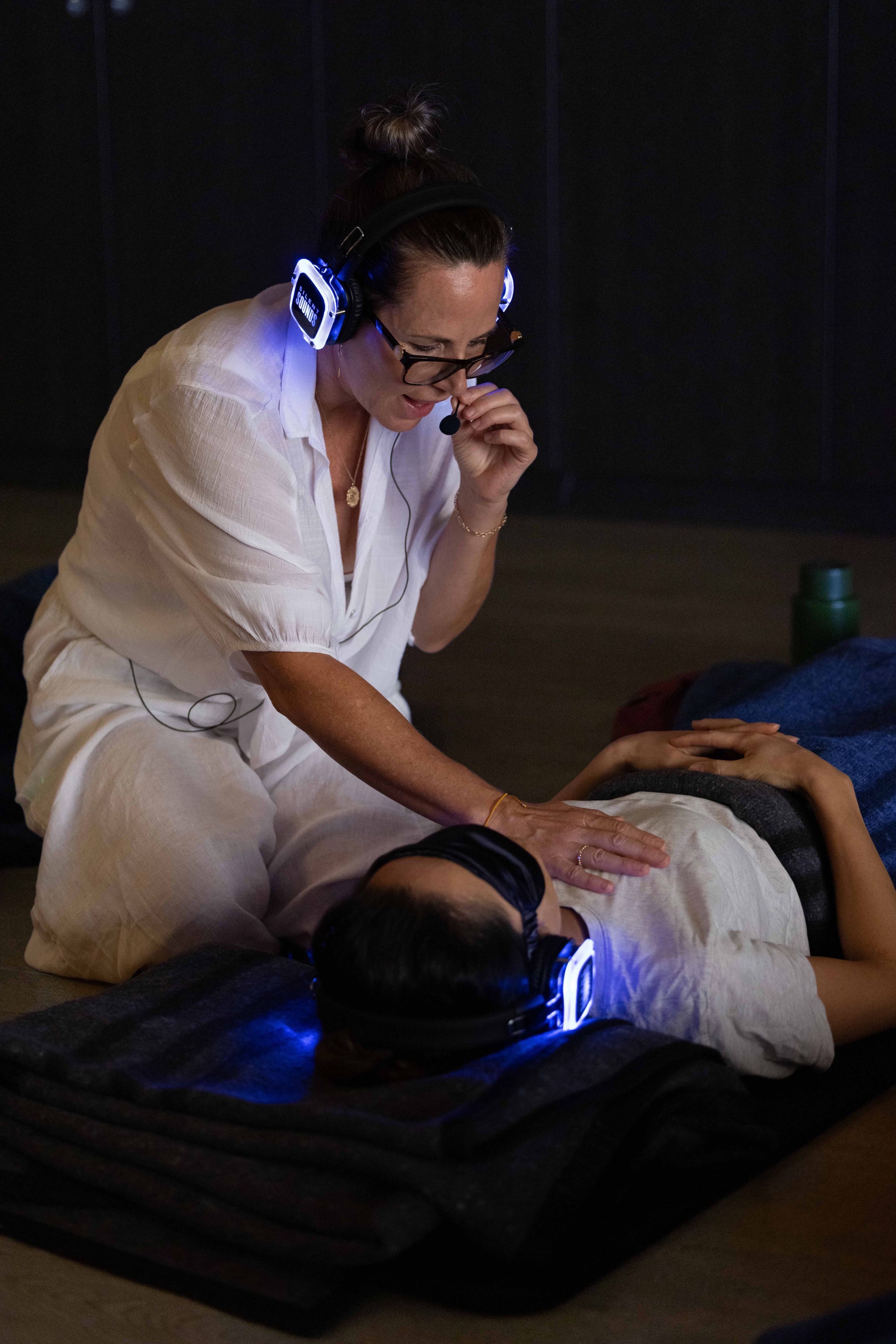 Brooke Stevenson kneeling beside a participant during a 9D breathwork session, both wearing LED headphones in a darkened studio with blue light