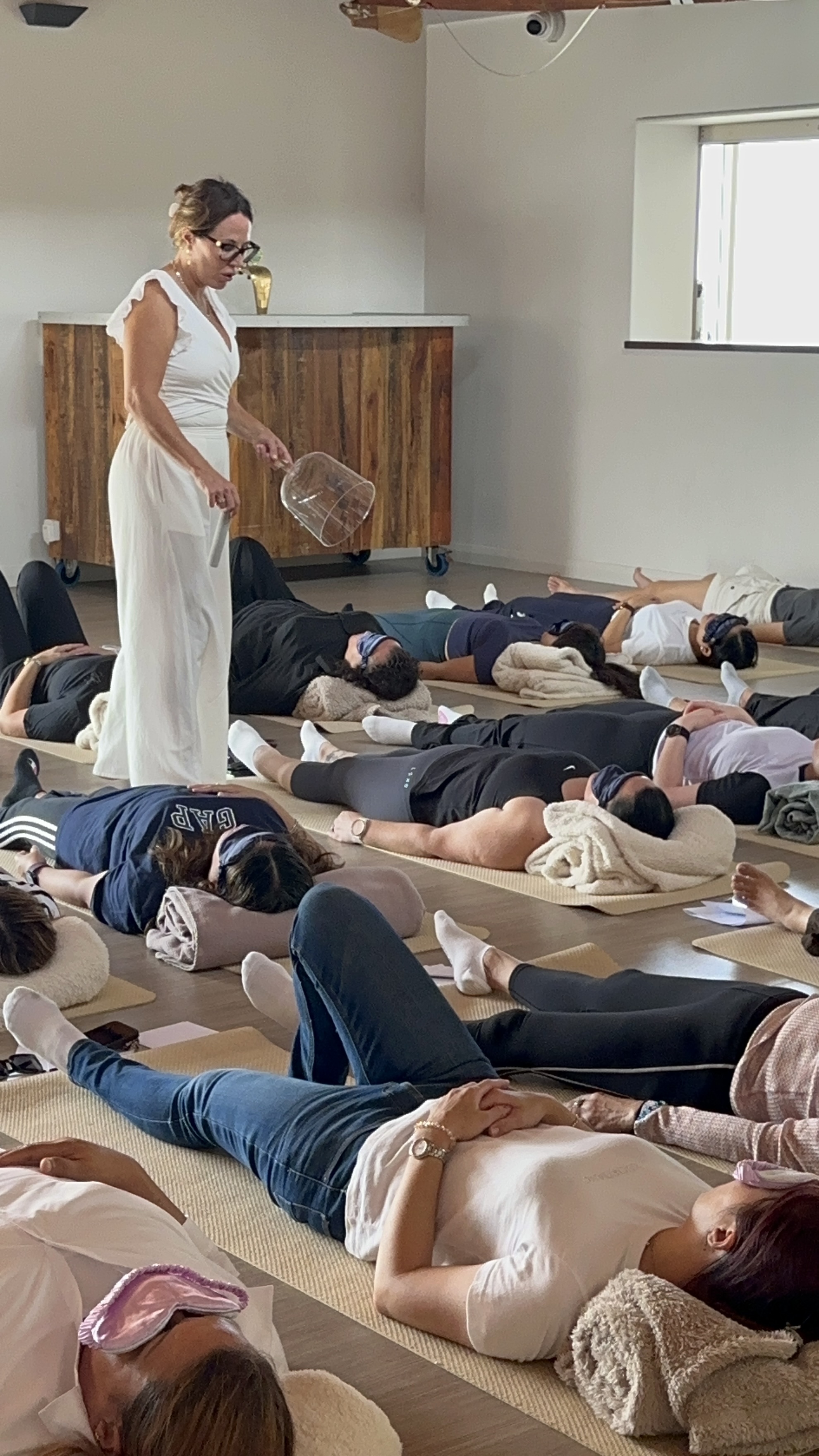 Brooke Stevenson leading a group breathwork session, walking among participants lying on mats with eye masks in a sunlit studio