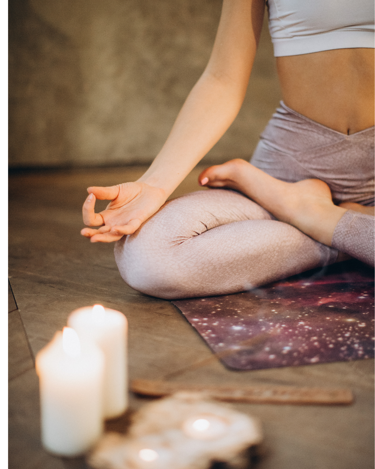 A woman seated cross-legged on a galaxy-print yoga mat in a candlelit room, one hand resting in a mudra position with candles and crystals in the foreground