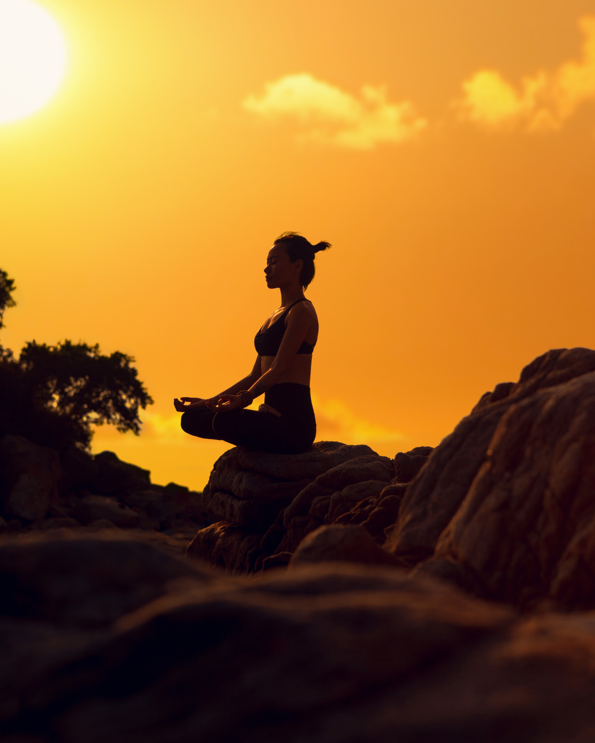 A figure sitting in meditation on coastal rocks at golden hour, silhouetted against a glowing sunset sky