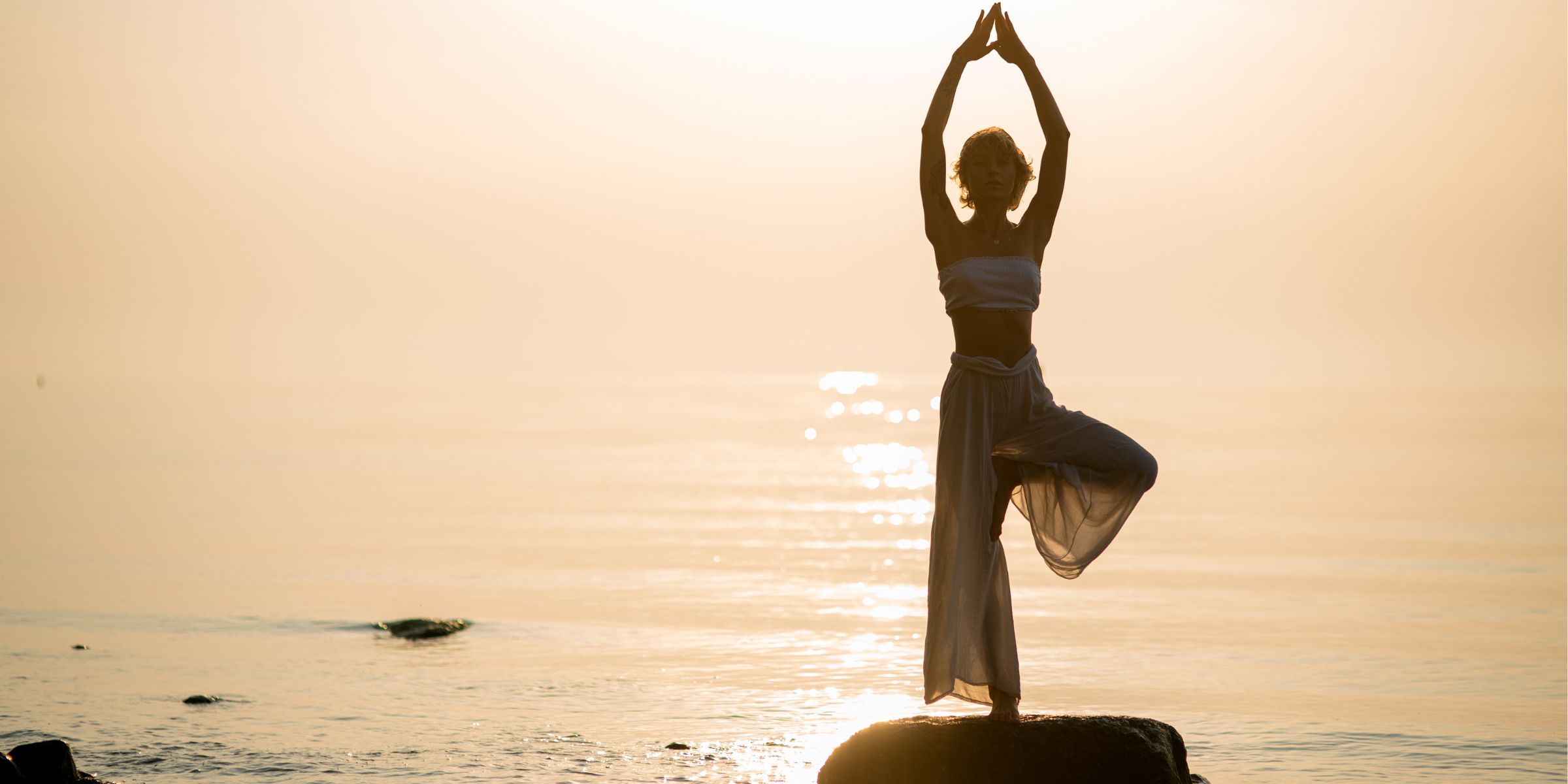 A woman practising yoga tree pose on rocks by the ocean at sunset, arms raised overhead against a warm golden sky