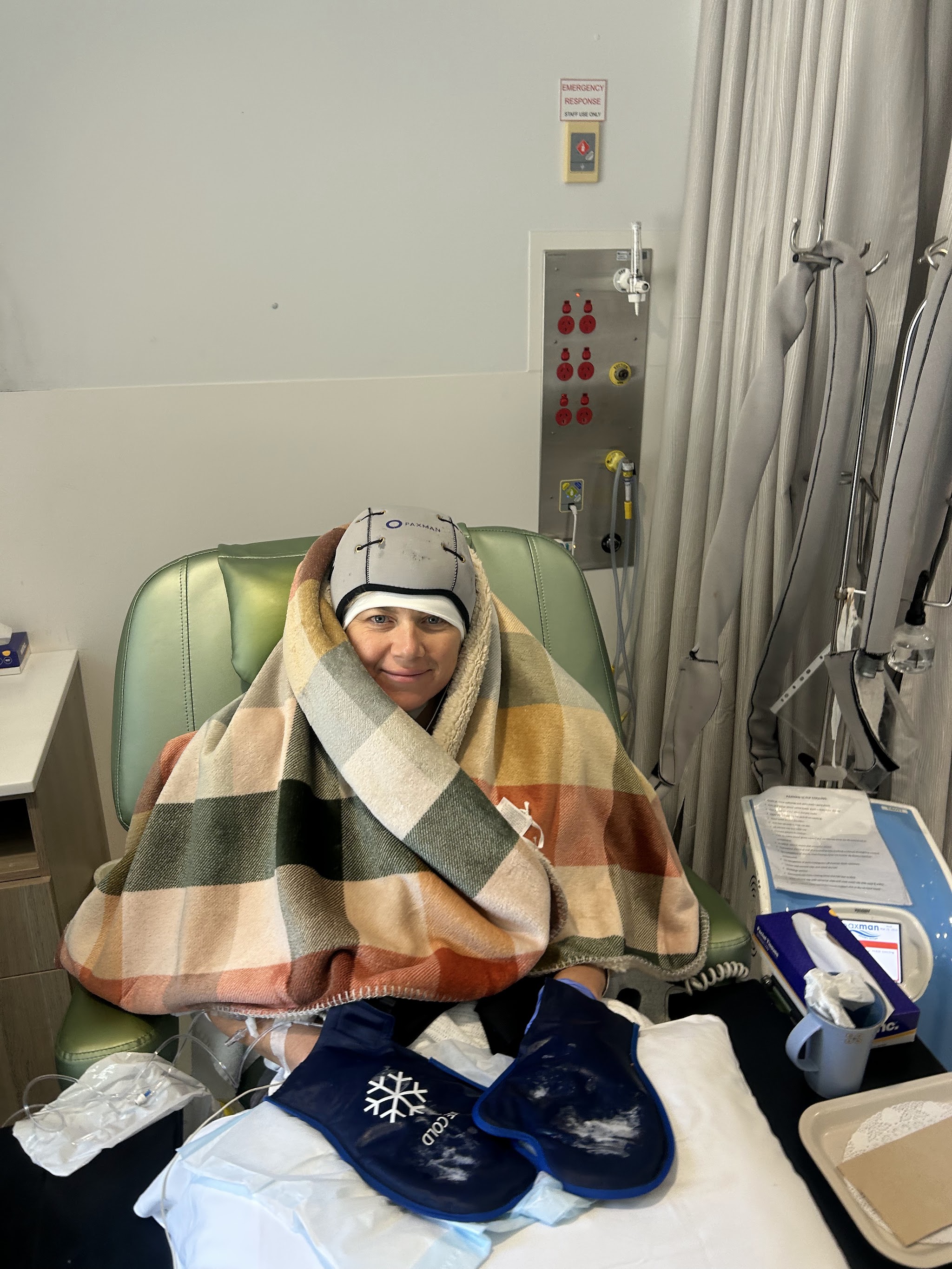 Colette during chemo wrapped in a plaid blanket with cold cap and gloves to protect her nails
