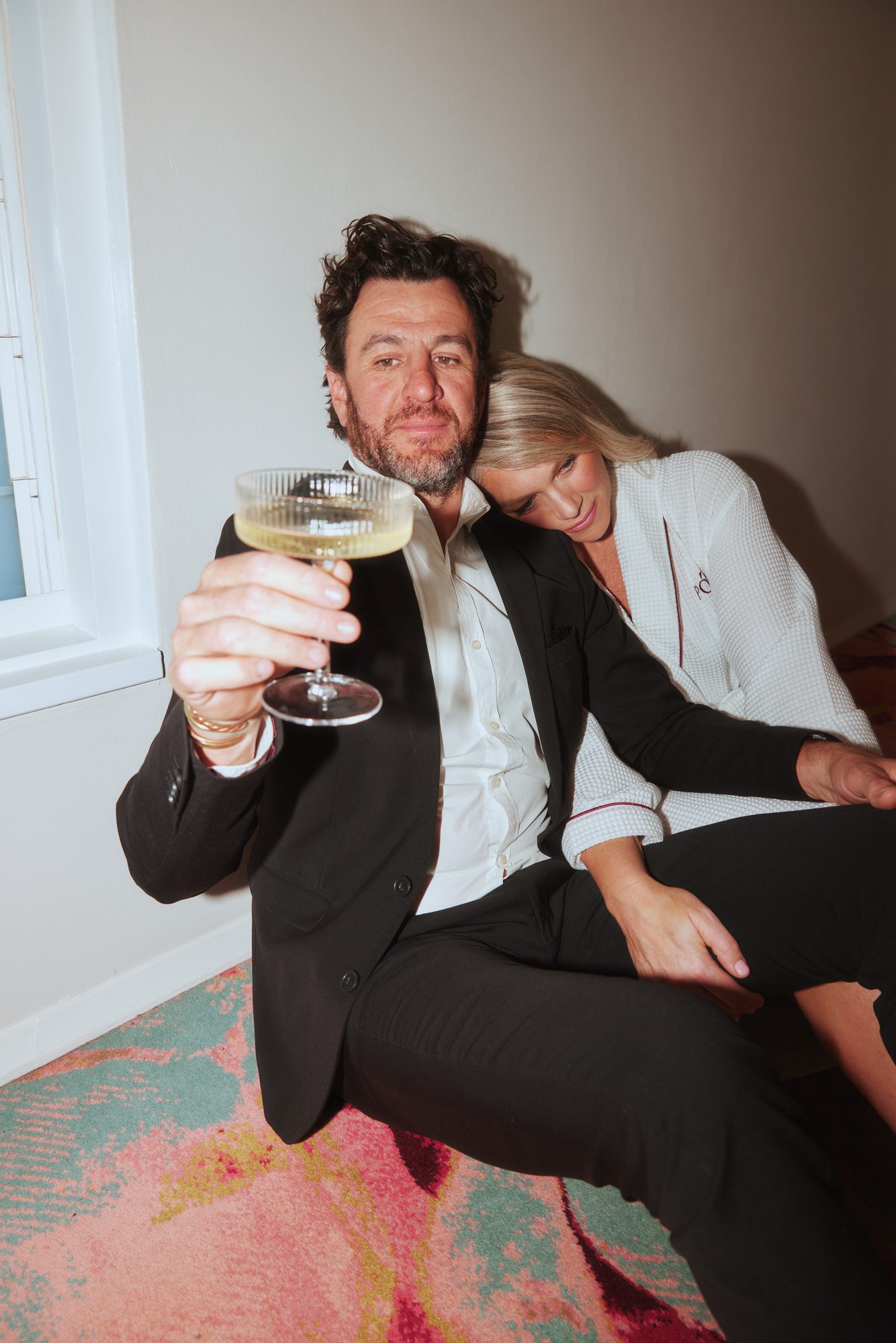 Colette Harvey in a white robe leaning on Joe, who holds a coupe glass
