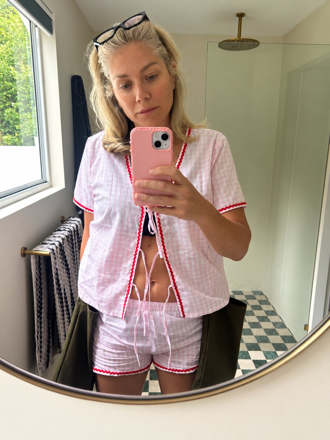 Colette Harvey taking a mirror selfie in a bathroom, in pink gingham pyjamas with a pink phone and glasses on her head