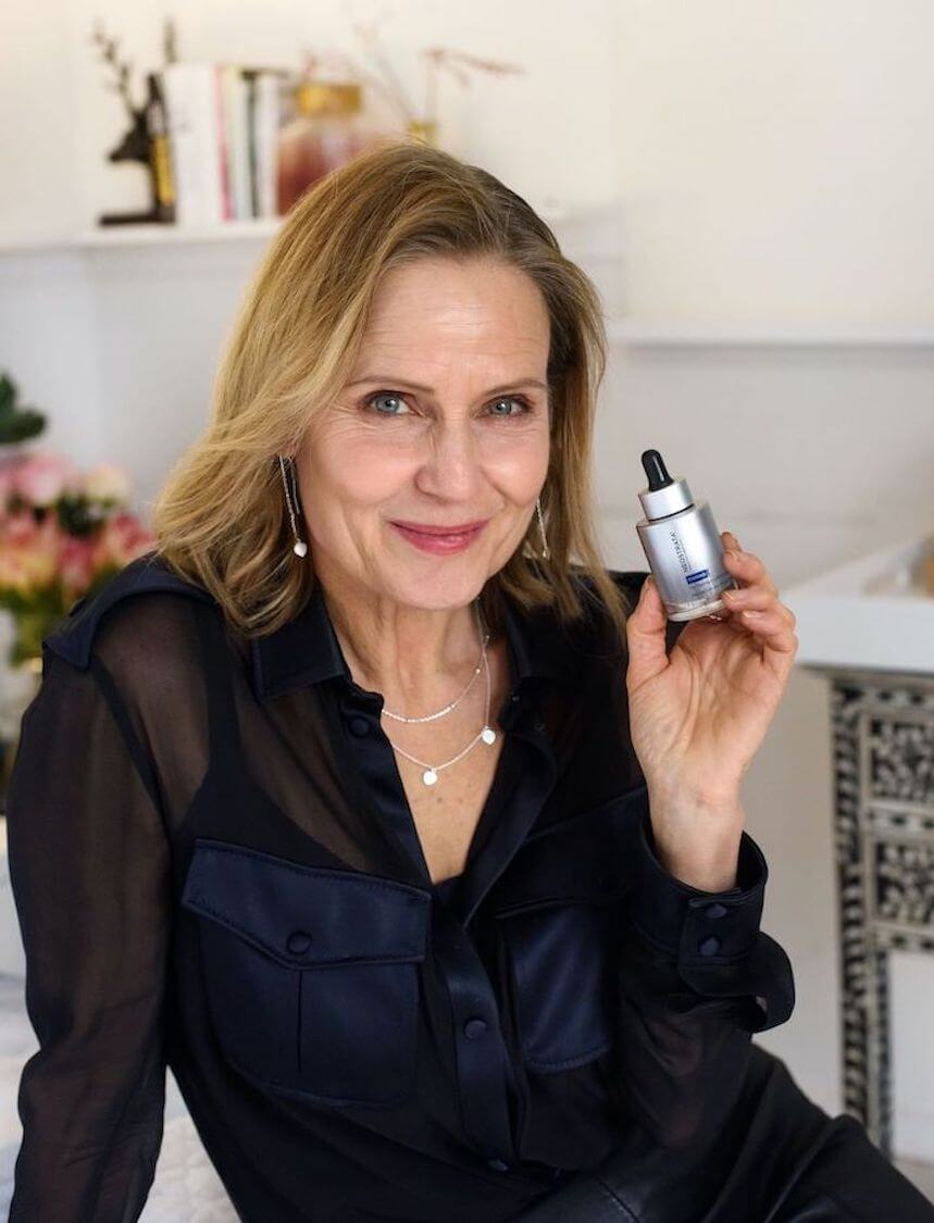 Shaynna Blaze in a navy silk blouse holding a Neostrata serum bottle in her bright living room