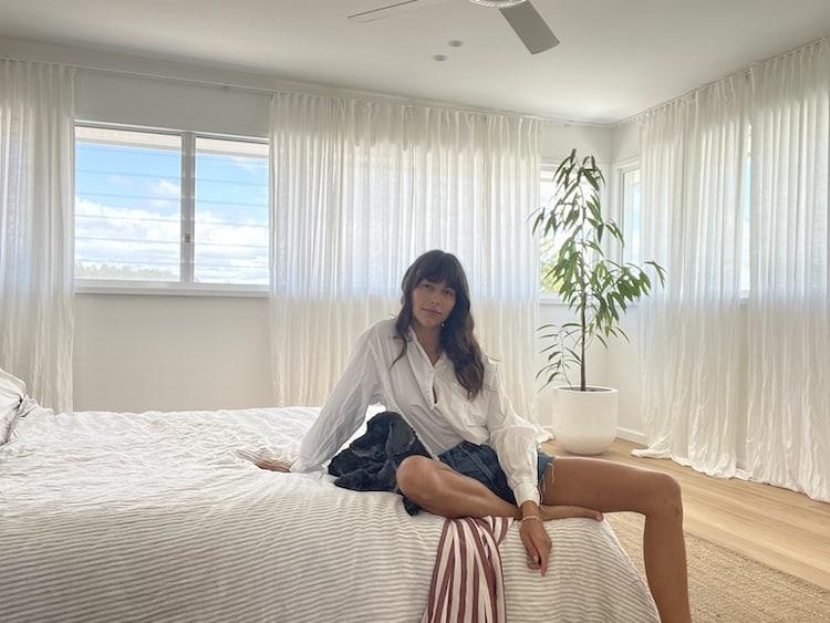 Helena Vestergaard seated on a white bed in a white shirt and denim skirt, light-filled bedroom
