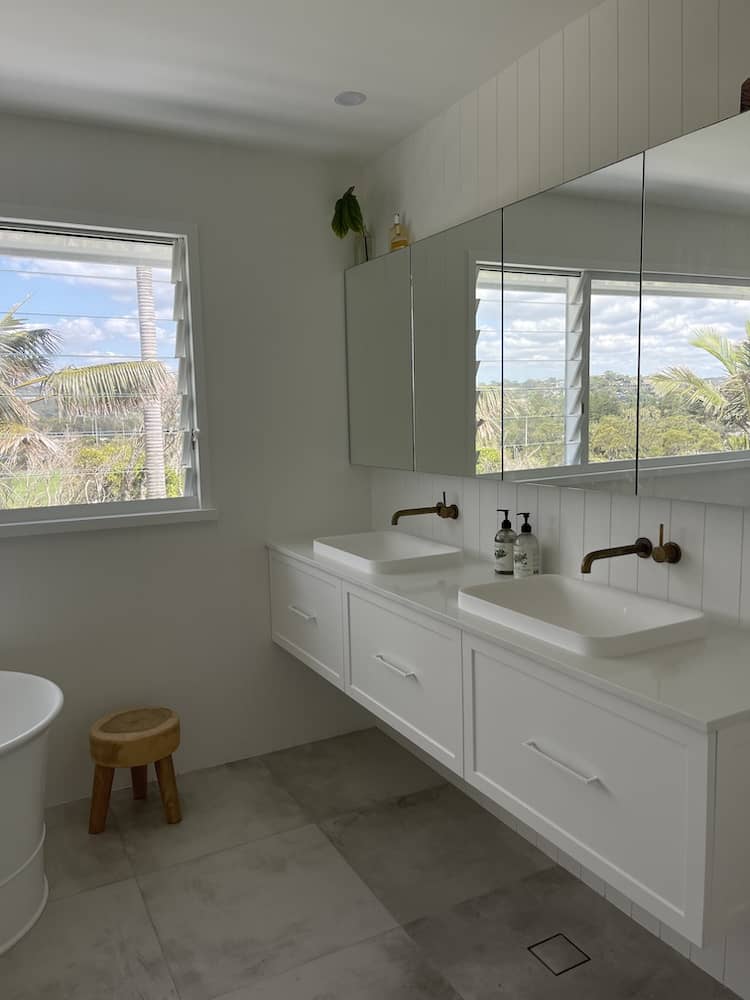 White coastal bathroom with double floating vanity, brass tapware, mirrored cabinets and palm-tree views