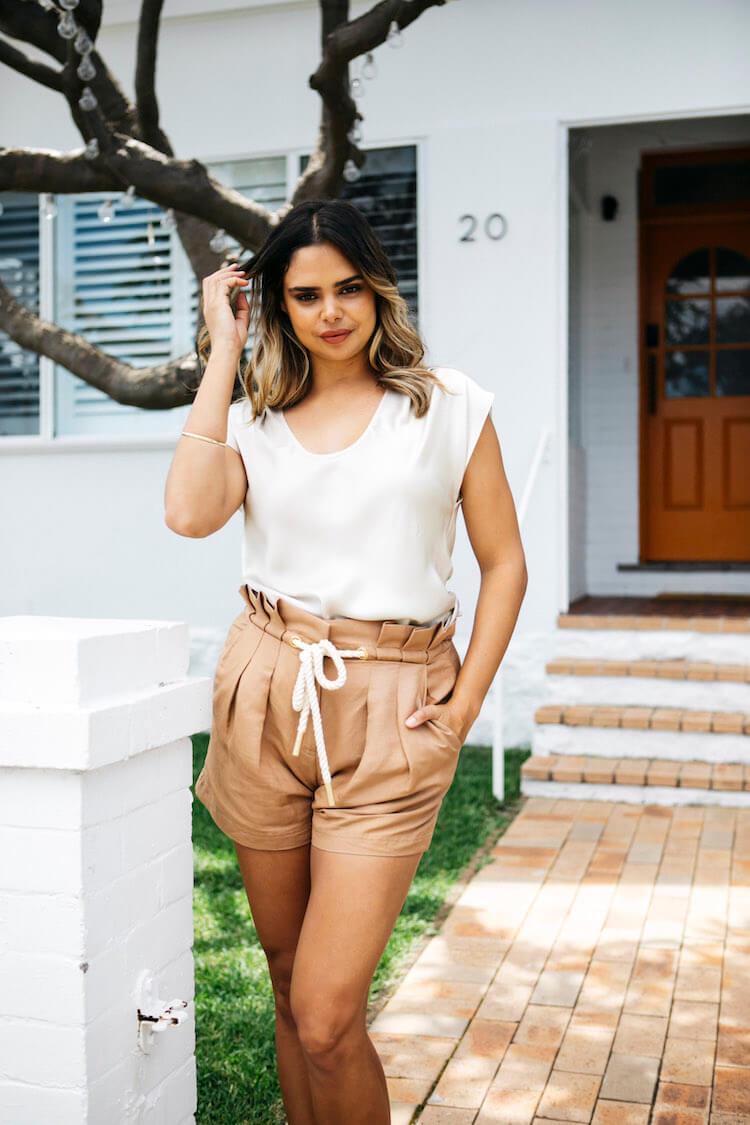 Samantha Harris in a white silk top and tan paperbag shorts standing outside her white weatherboard home