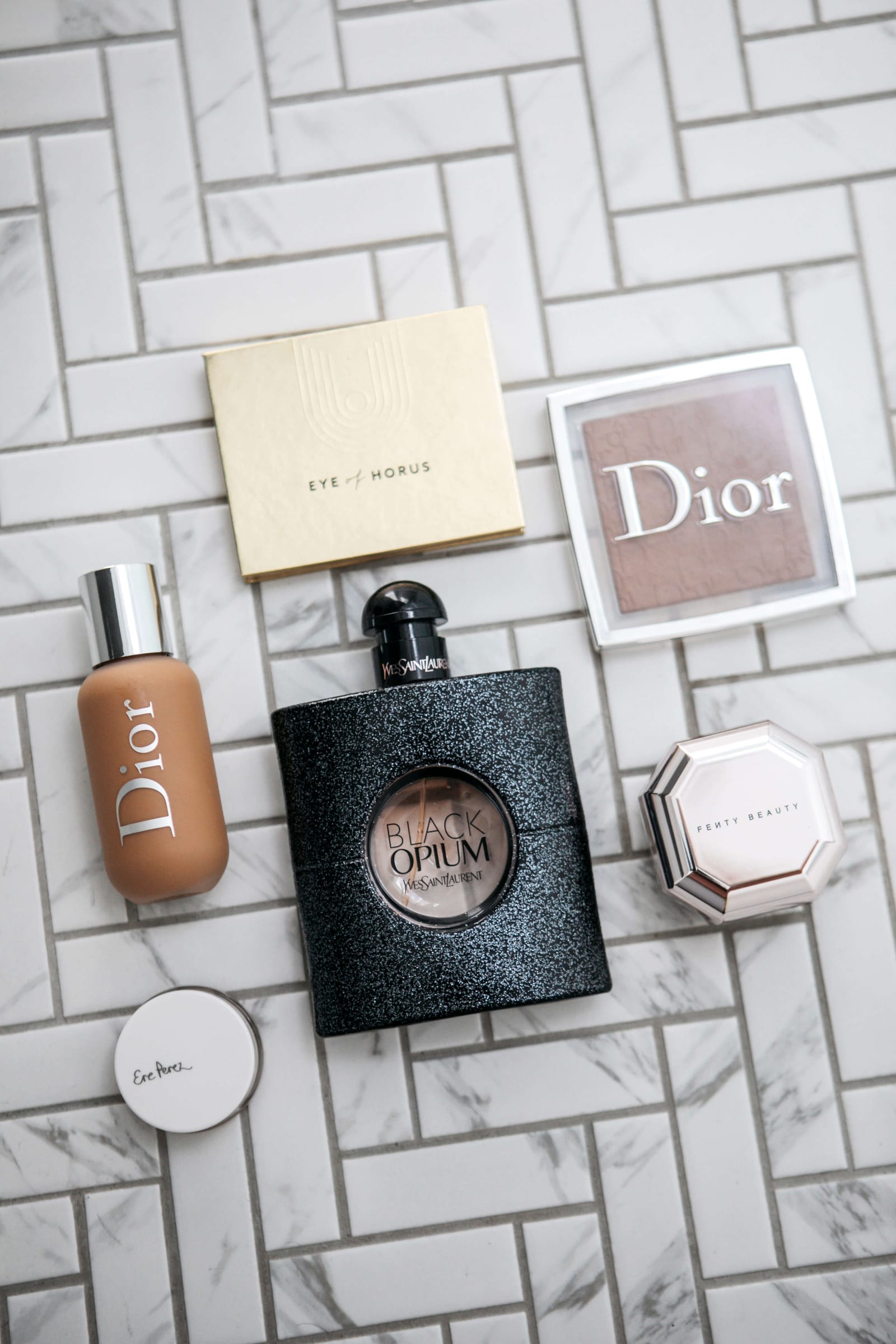 Flat lay of Dior foundation, YSL Black Opium perfume, Dior highlighter, Fenty Beauty and Eye of Horus on tile