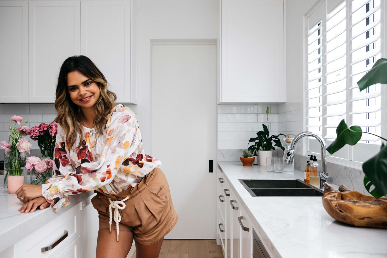 Samantha Harris in a floral blouse and tan shorts leaning on the kitchen bench with pink flowers nearby