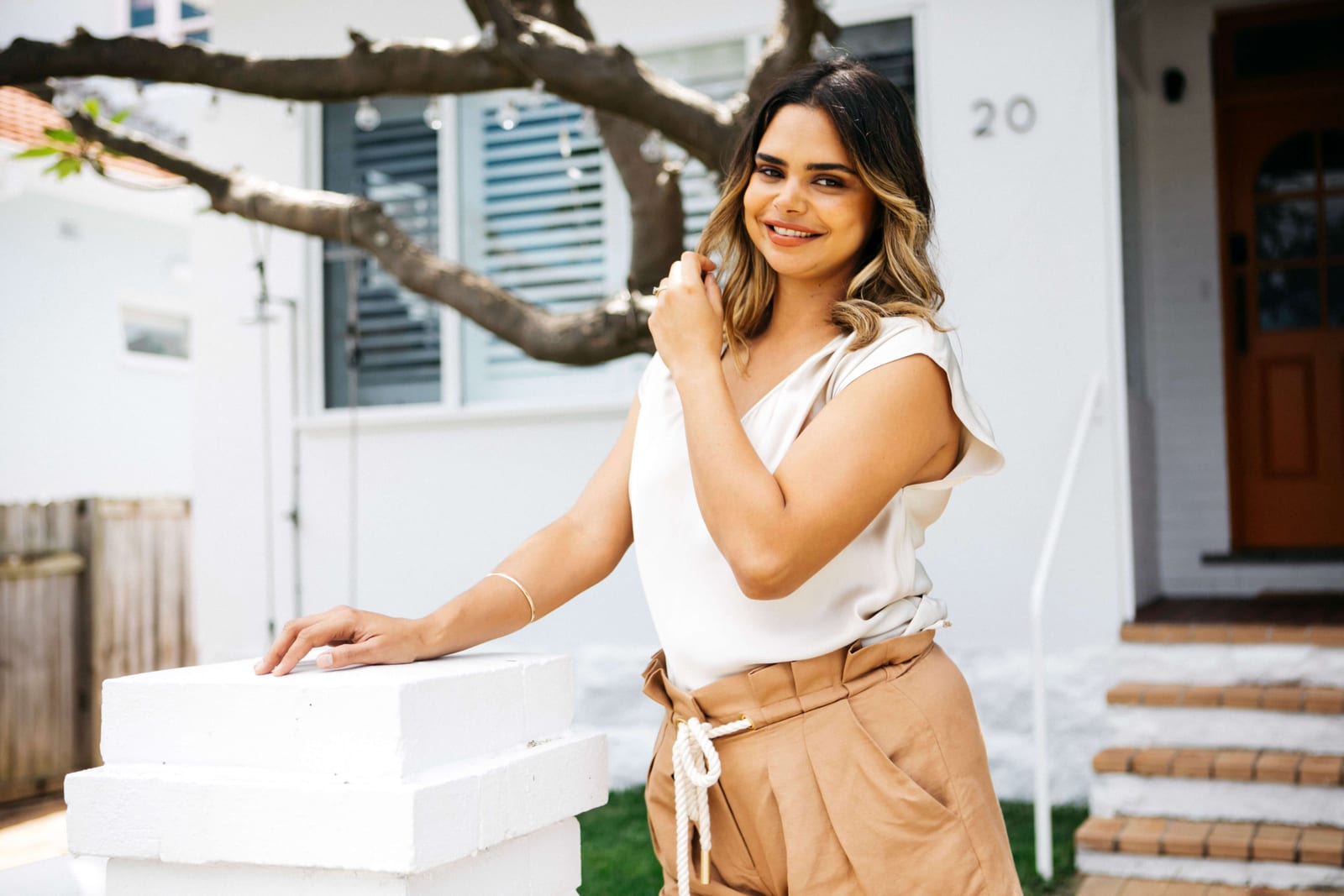 Samantha Harris smiling and leaning on a white pillar outside her home in a cream silk top and tan shorts