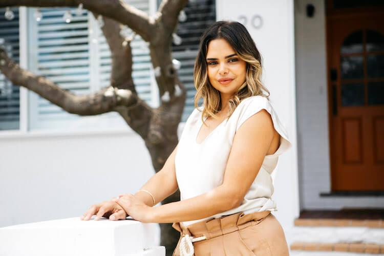 Close-up of Samantha Harris leaning on a white pillar in a cream top, smiling softly with ombre waves