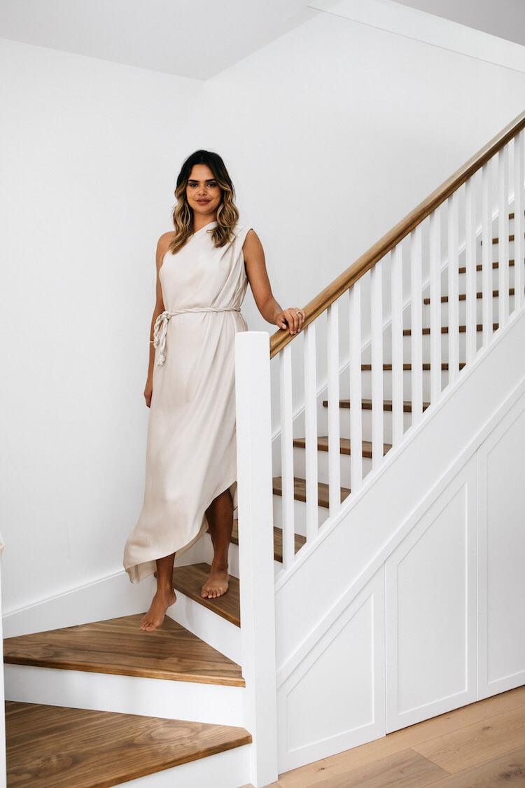 Samantha Harris in a cream one-shoulder draped dress standing on timber stairs with a white banister