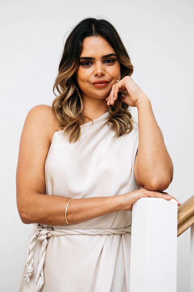 Portrait of Samantha Harris in a cream one-shoulder dress, chin resting on her hand at the stair railing