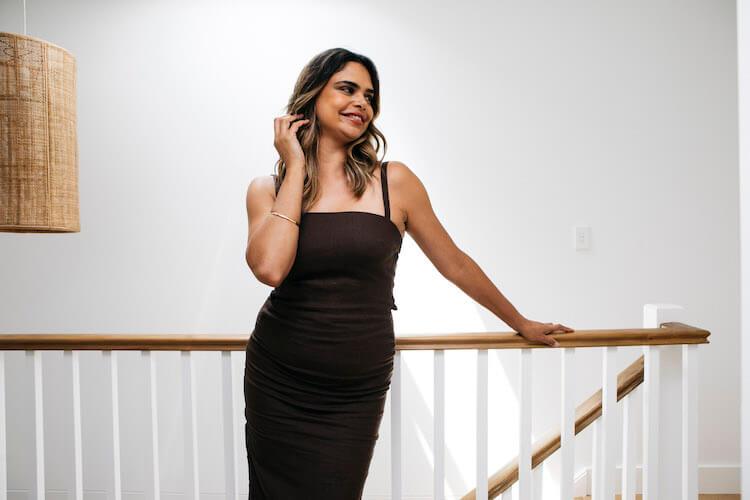 Samantha Harris in a fitted black dress leaning on a staircase railing beside a rattan pendant lamp