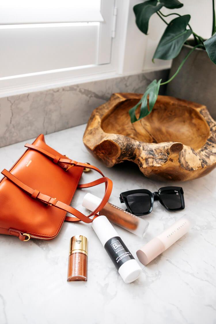 Tan leather handbag, Fenty Beauty products, Biologi serum, sunglasses and a wooden bowl on marble