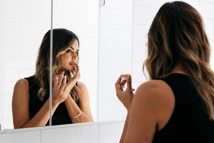Samantha Harris applying skincare in her bathroom mirror, wearing a black sleeveless top