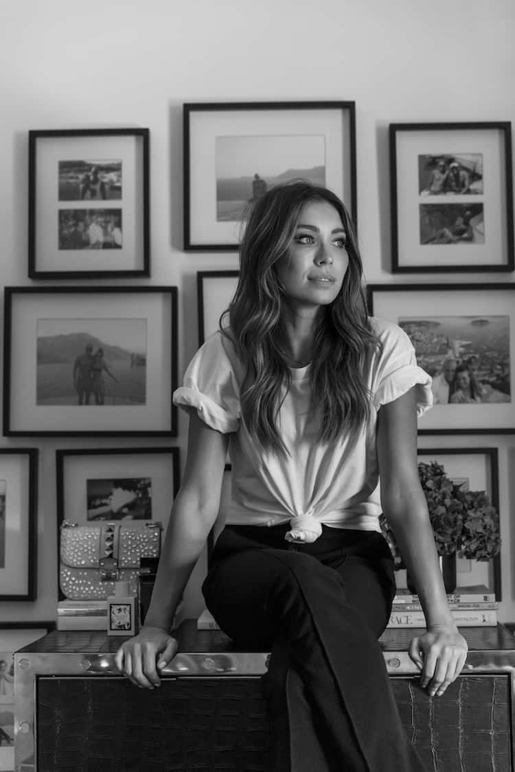 Black-and-white portrait of Rozalia seated on a credenza in a knotted white tee, gallery wall behind