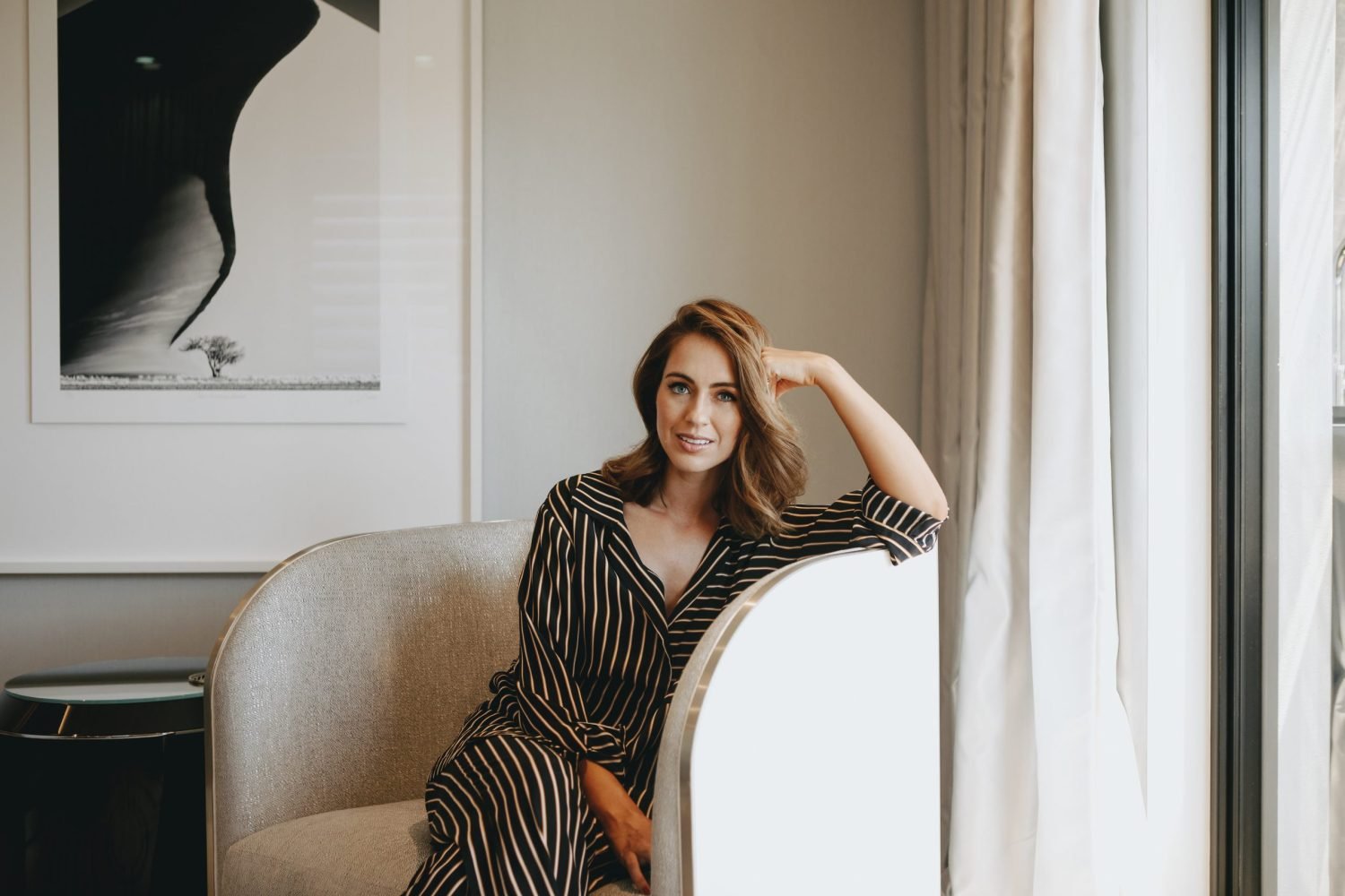Woman with short dark hair in a black and white robe sitting on a beige sofa in a clinic waiting area, smiling softly.