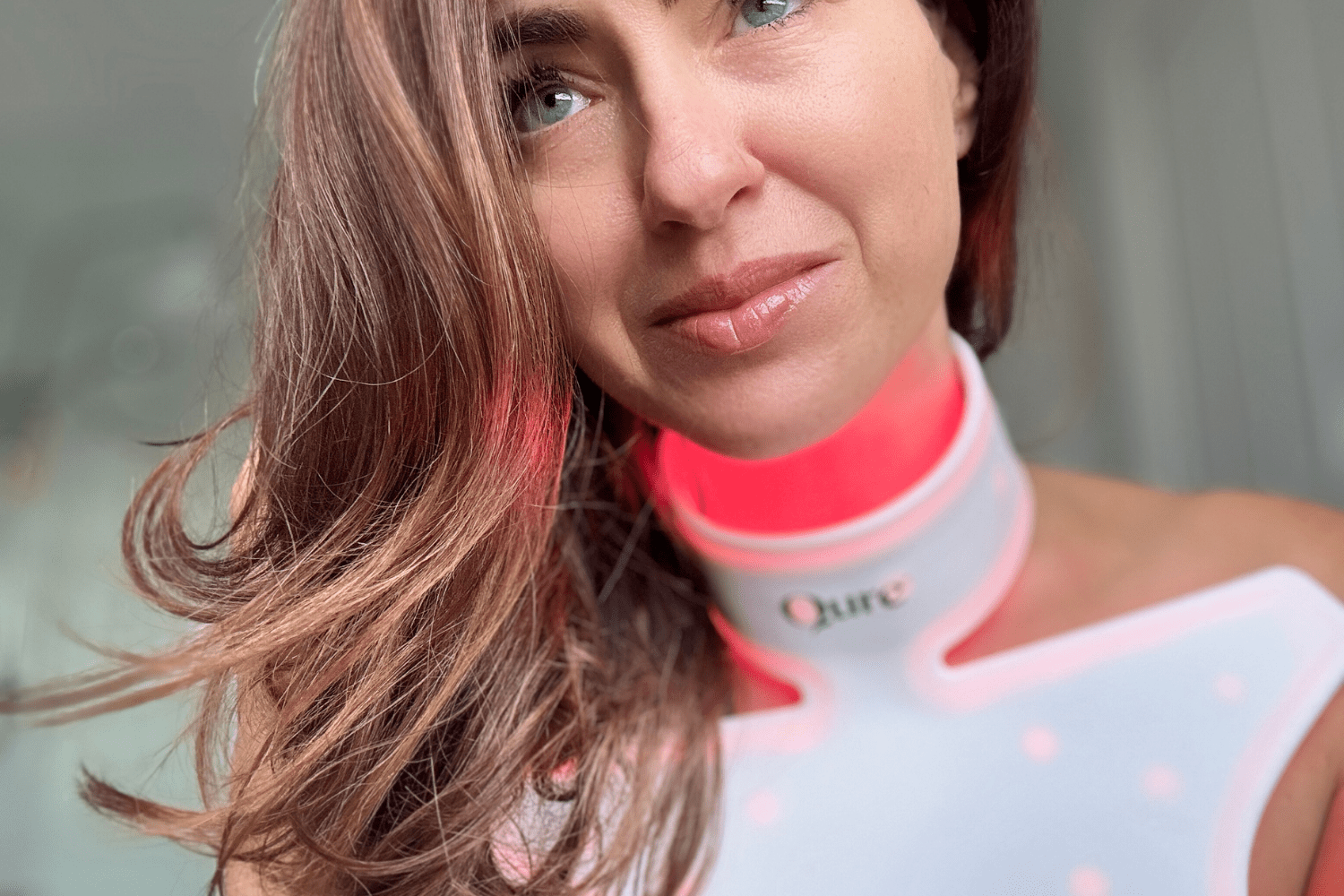 Soft-focus portrait of woman using Qure Neck & Décolletage LED Mask, red light highlighting neck and chest.